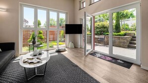 Een moderne woonkamer met een grote schuifpui naar een tuin, een flatscreen televisie, een grijze bank en een salontafel.