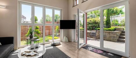 A modern living room with a large sliding glass door leading to a garden, a flat-screen TV, a grey sofa, and a coffee table.