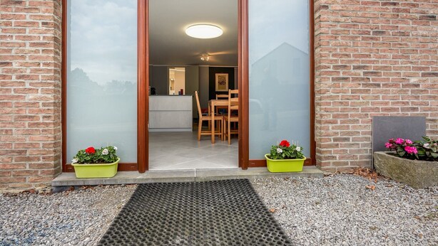 A brick building with a sliding glass door entrance, a wooden dining table and chairs inside, and two flower pots with pink flowers outside. - Cottage in Aywaille with Garden (Aywaille)