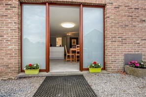 Un bâtiment en briques avec une entrée dotée d’une porte-fenêtre coulissante, une table et des chaises en bois à l’intérieur, et deux jardinières avec des fleurs roses à l’extérieur.