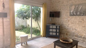 A room with a stone wall, a television mounted above a cabinet, a sliding glass door leading to a garden, and a small table with a basket. - ELOISE             . (Sainte-Marie-de-Ré)