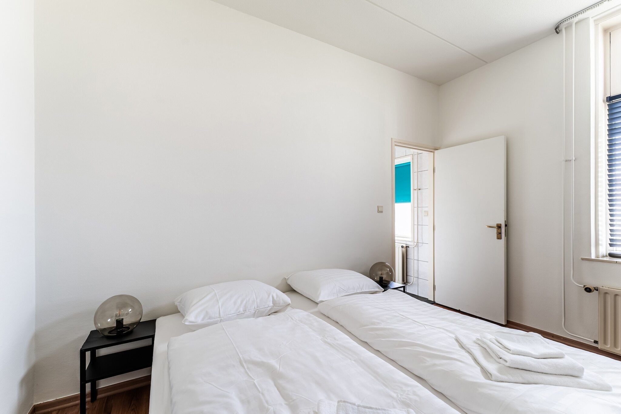 Un dormitorio minimalista con dos camas, una mesita de noche y una puerta que da a otra habitación.