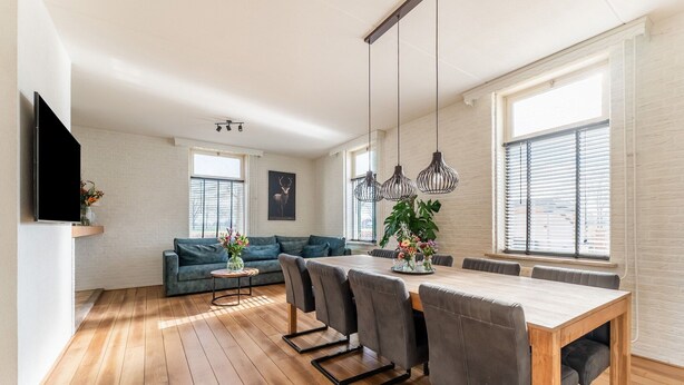 A modern living room with a wooden floor, a sofa, a dining table with chairs, pendant lights, and a TV mounted on the wall. - Farmhouse in Silvolde with Hot Tub (Silvolde)