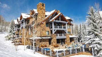 Un albergue de varios pisos con construcción de piedra y madera, un telesilla y árboles cubiertos de nieve.