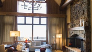 A spacious living room with a high ceiling, a stone fireplace, and a large window providing a view of the outdoors.