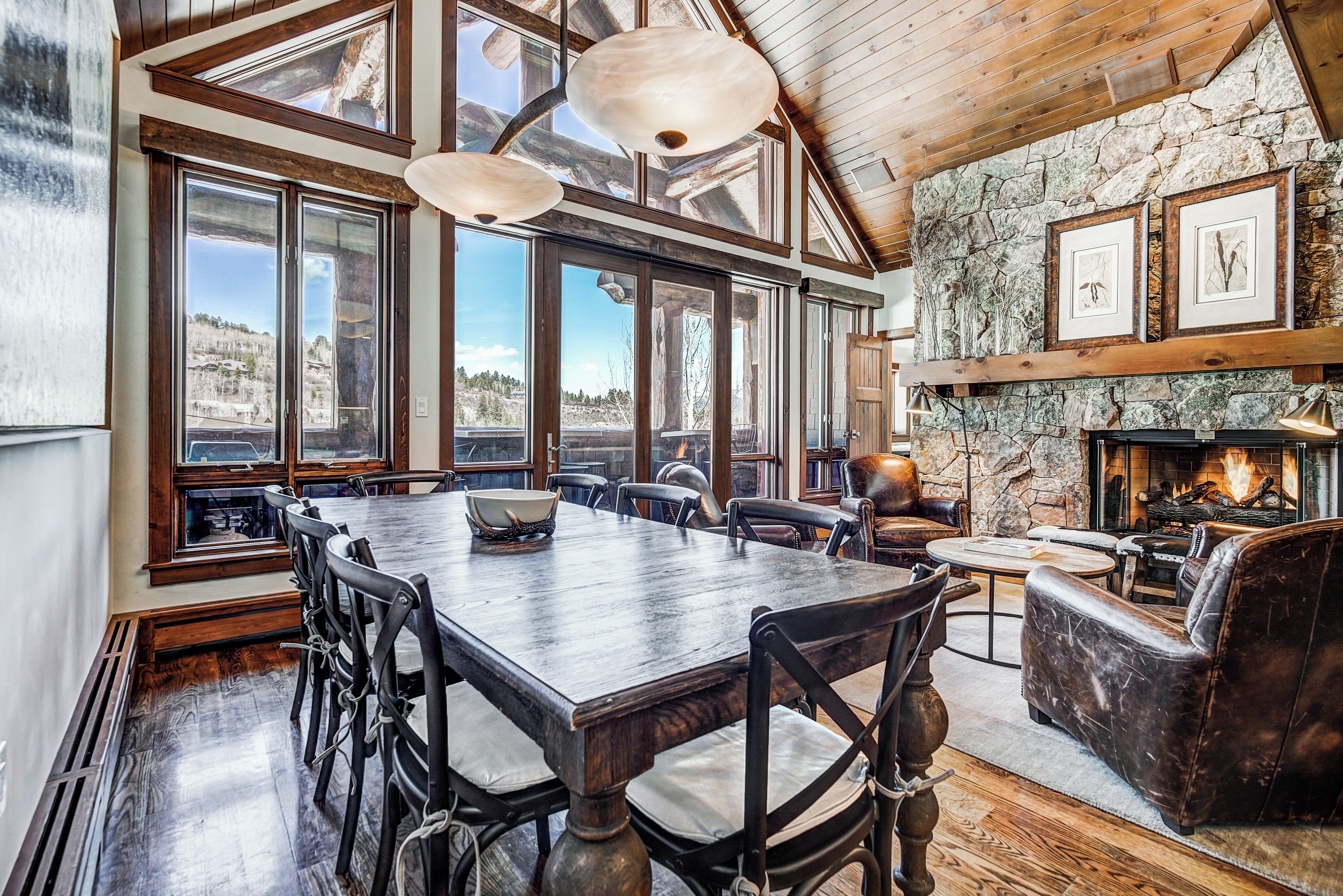 Un comedor amplio con una gran mesa de madera y sillas, ventanas grandes con vistas al exterior y una chimenea de piedra con obras de arte enmarcadas arriba.