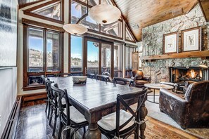 A spacious dining area with a large wooden table and chairs, large windows offering a view of the outdoors, and a stone fireplace with framed artwork above. - Bachelor Gulch Village (Avon)