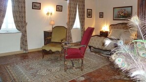 A traditional room with wooden beams, a patterned rug, a red chair, a bed with a floral painting, and a peacock feather decoration.