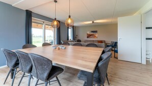 En rummelig spiseplads med et stort træbord og moderne stole, hængende pendellamper og udsigt til det fri gennem store vinduer.