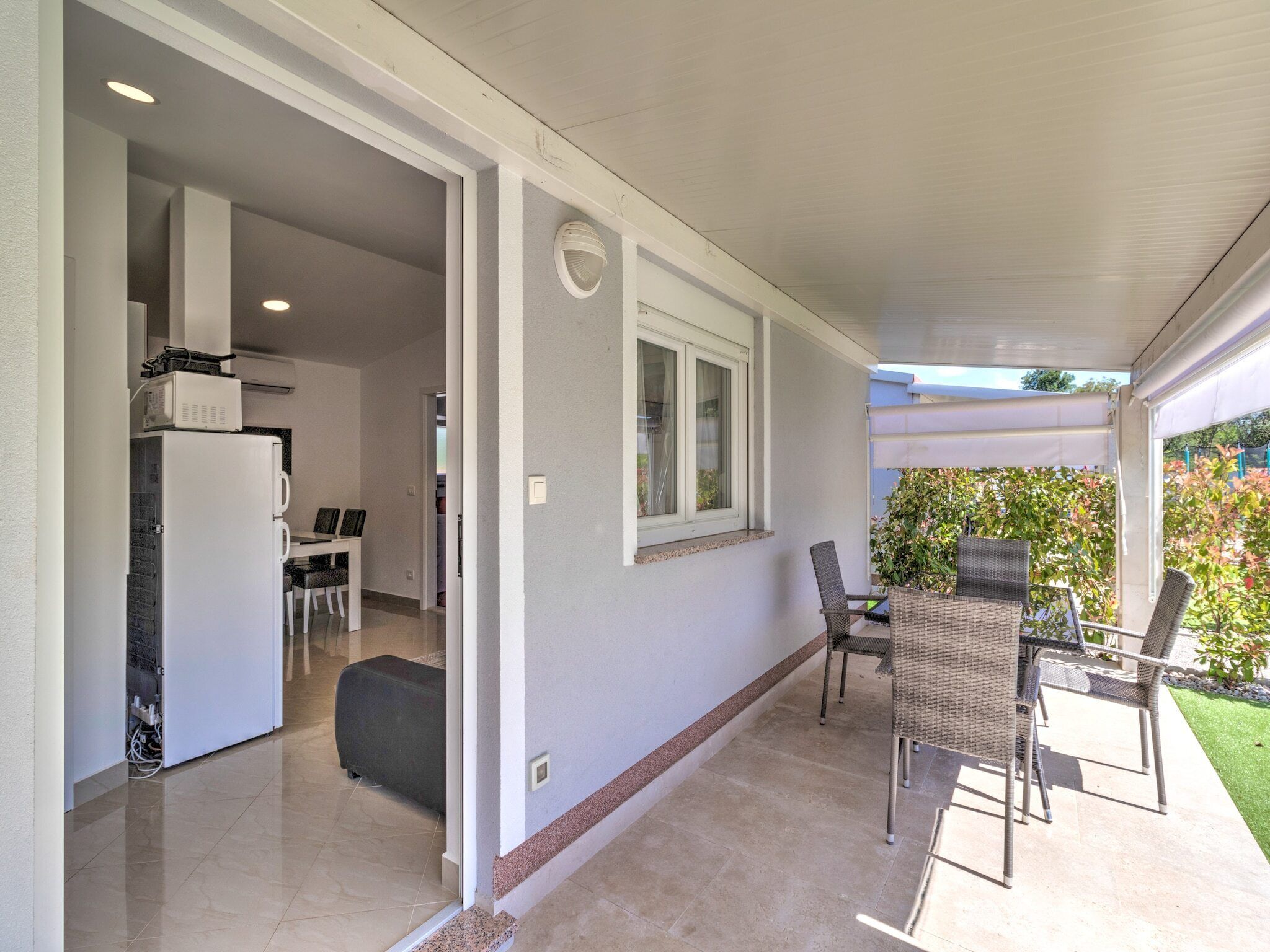 A covered patio with outdoor seating, a kitchen area with a refrigerator and microwave, and a view of a garden.