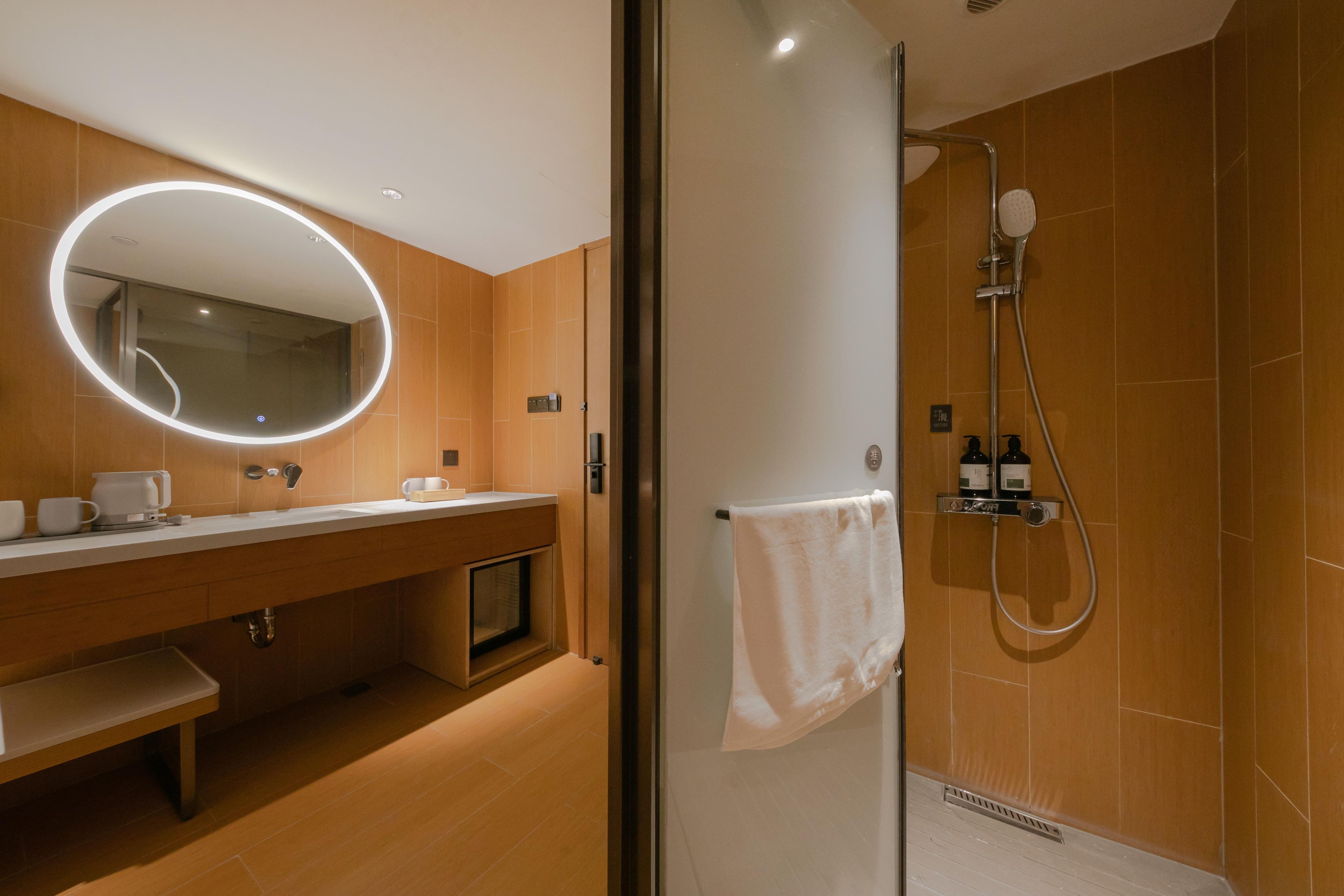 Une salle de bain moderne dotée d’une douche à parois de verre, d’un grand miroir rond avec éclairage intégré et d’un meuble-lavabo en bois avec un banc.