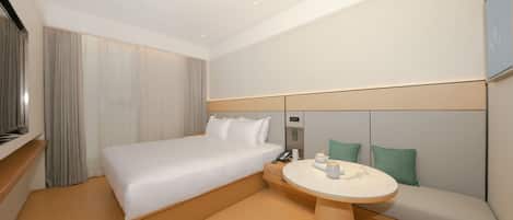 Una habitación de hotel moderna con una cama grande, una mesita de noche con un teléfono y una taza, una pequeña mesa redonda con un hervidor y un sofá con cojines.