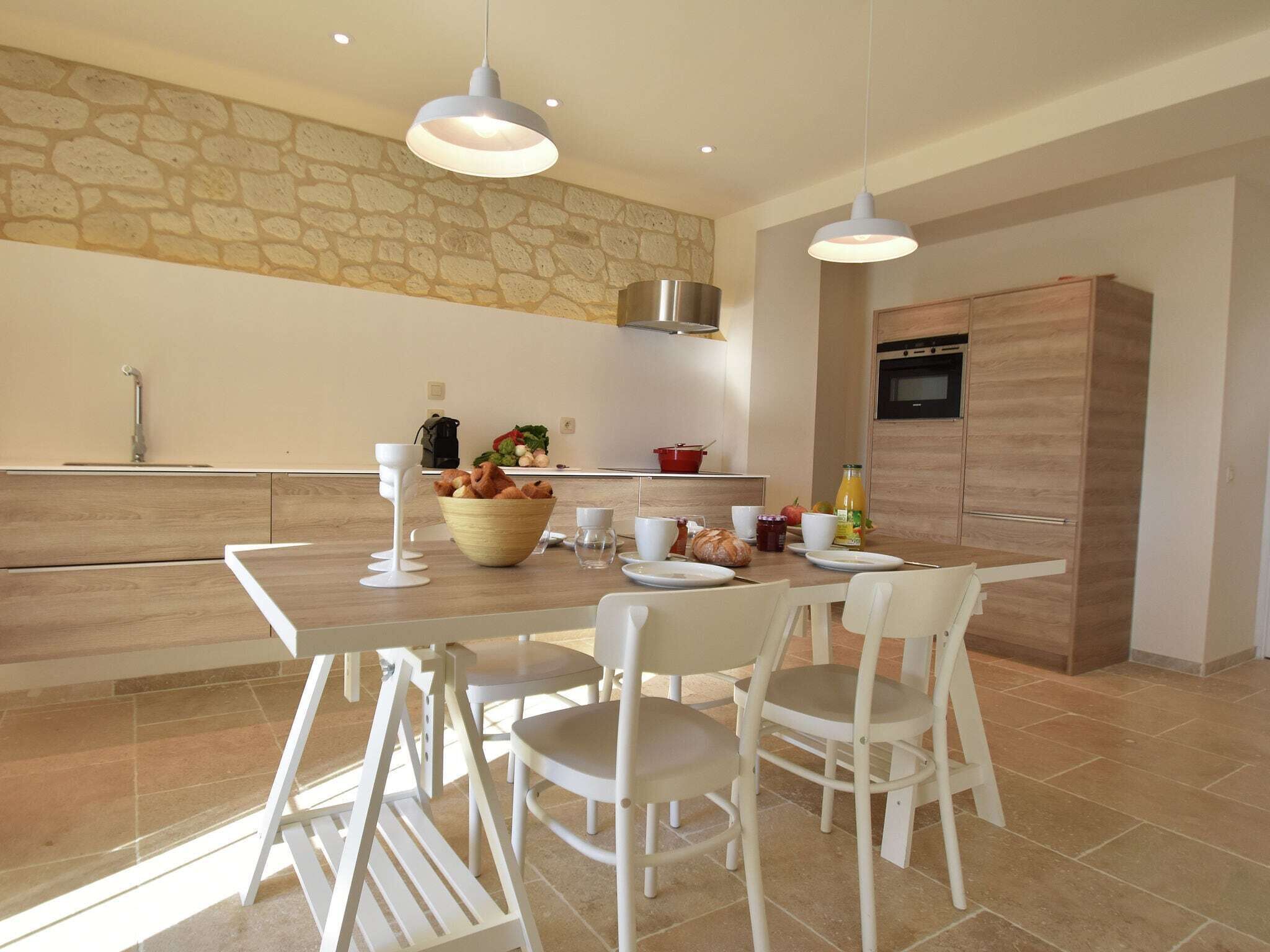A modern kitchen with a stone wall, a wooden dining table, white chairs, and a built-in oven.
