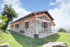 Un edificio de piedra con tejado inclinado y contraventanas de madera, rodeado de césped y un gran árbol.