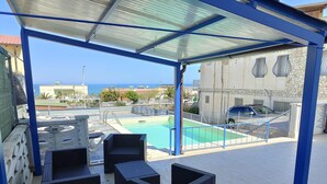 A covered outdoor area with a pool, black wicker furniture, and a view of the sea and buildings. - Alcamo Marina Villa | Pool (Alcamo Marina)
