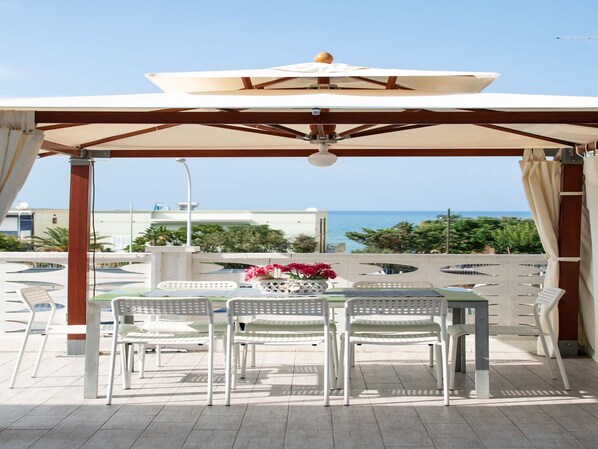 A covered outdoor seating area with a table and chairs, overlooking a pool and the sea.
