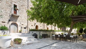 An outdoor courtyard with stone flooring, a seating area with tables and chairs, and a building with stone walls and arched windows.