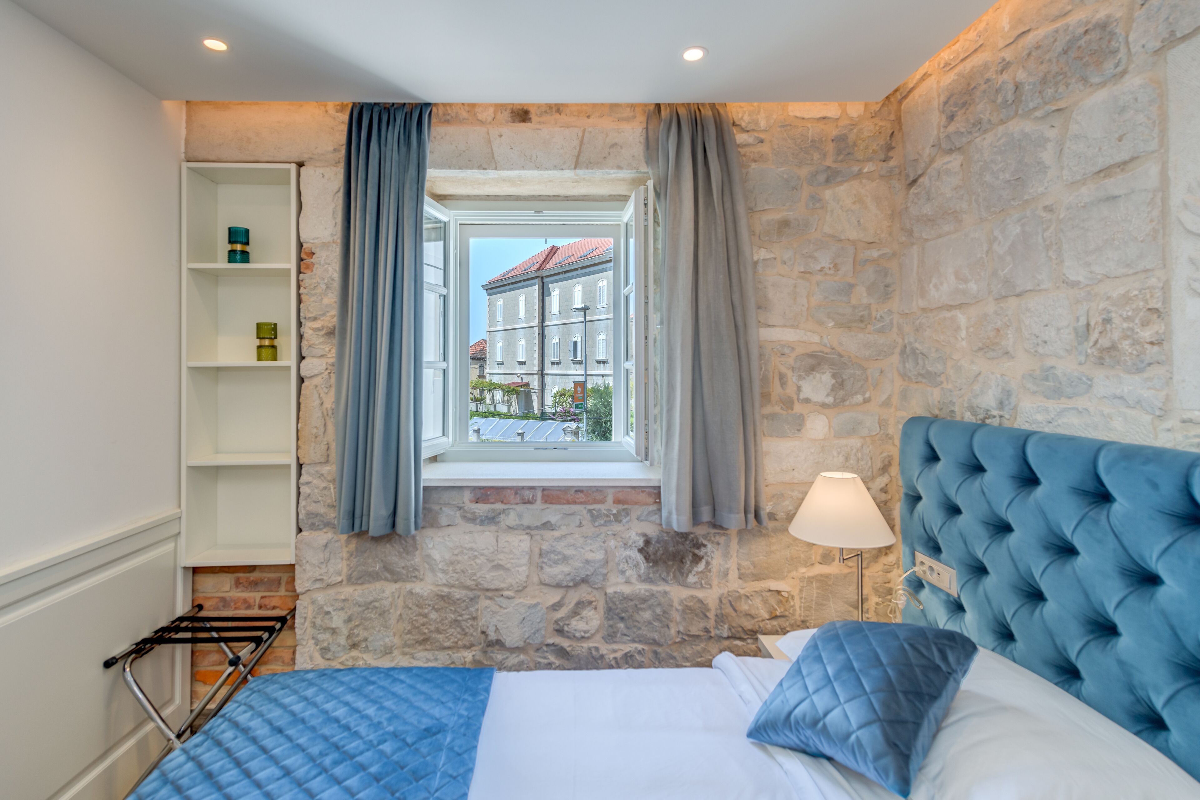 A bedroom with a stone wall, a window with curtains, a bed with blue bedding, and a bedside lamp.
