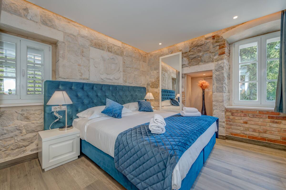 Un dormitorio moderno con una pared de piedra, una cama grande con ropa de cama azul, una mesita de noche con una lámpara y vistas a la vegetación exterior.