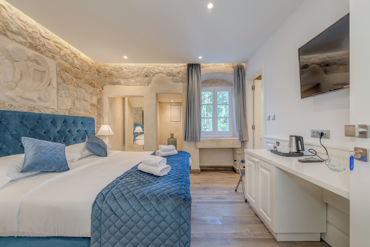 A hotel room with a stone wall, a bed with blue bedding, a small kitchenette, and a television mounted on the wall.