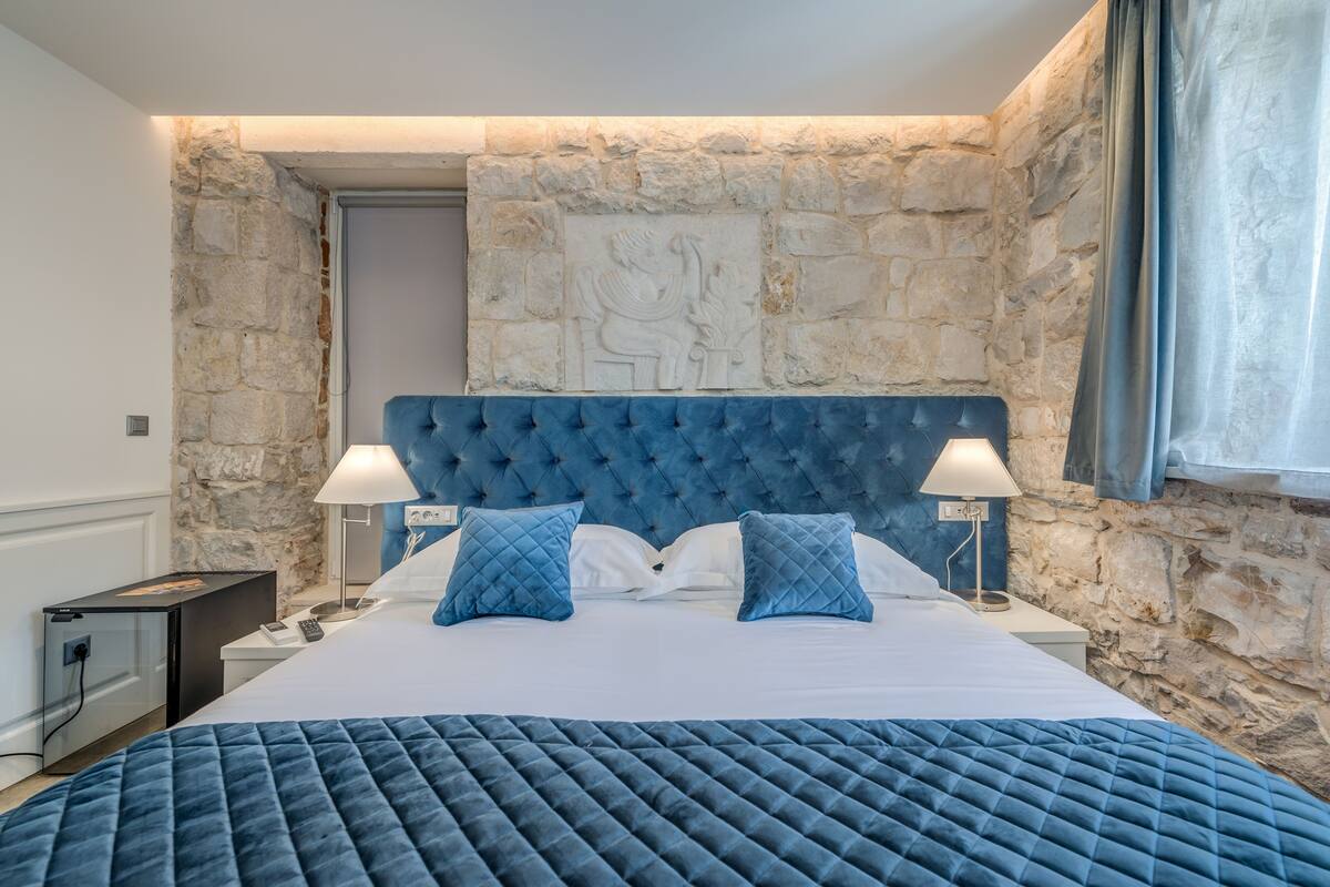 Un dormitorio con pared de piedra, cabecera capitonada, cama con ropa de cama azul, dos lámparas de noche y una mesita de noche con un teléfono y una lámpara.