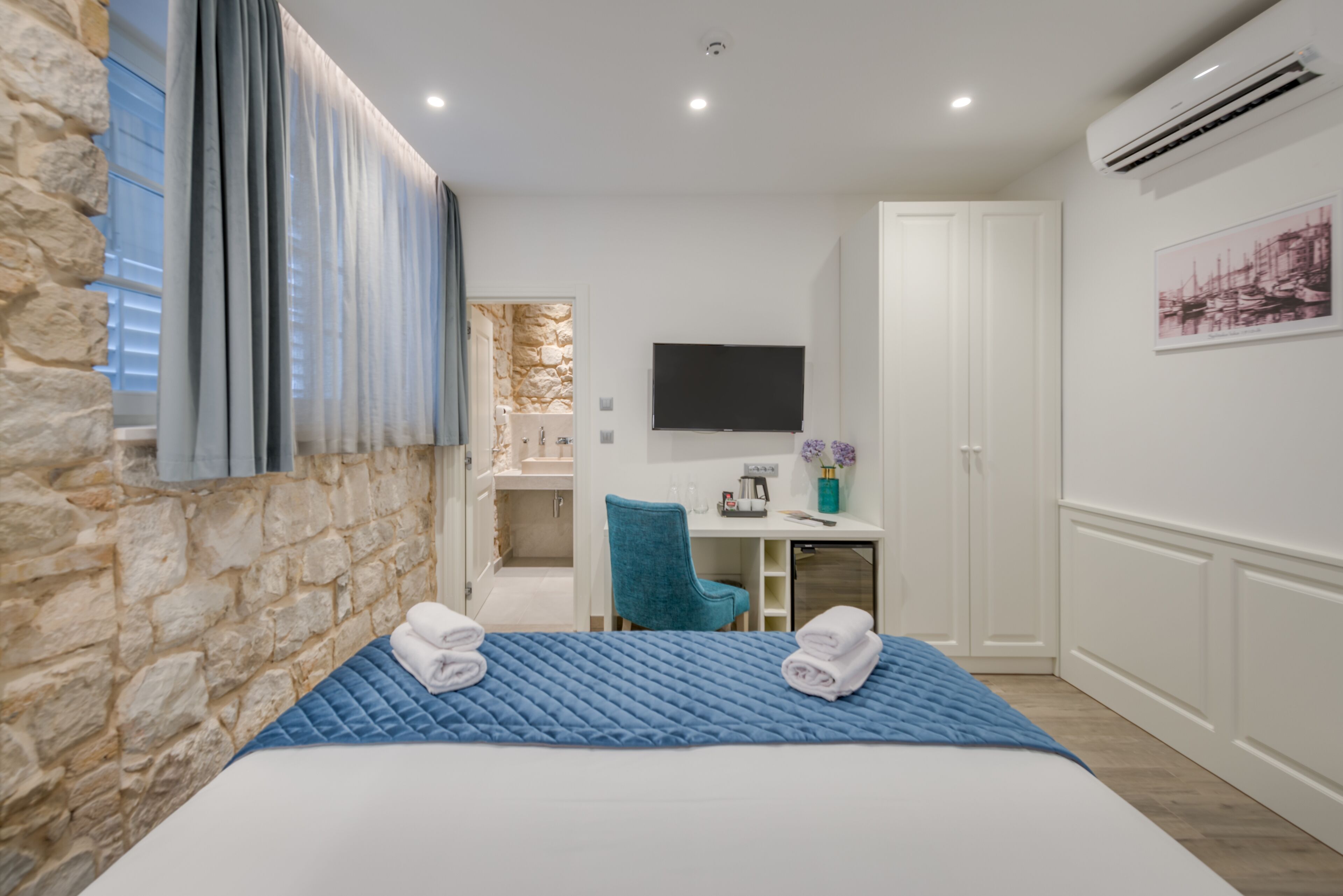 A hotel room with a stone wall, a bed with a blue quilt, a desk with a flat-screen TV, a chair, and a bathroom visible through an open door.