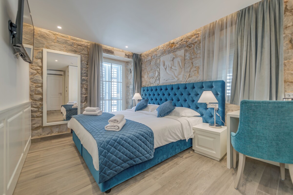 A bedroom with a stone wall, a large bed with a blue headboard, a bedside table with a lamp, and a chair.