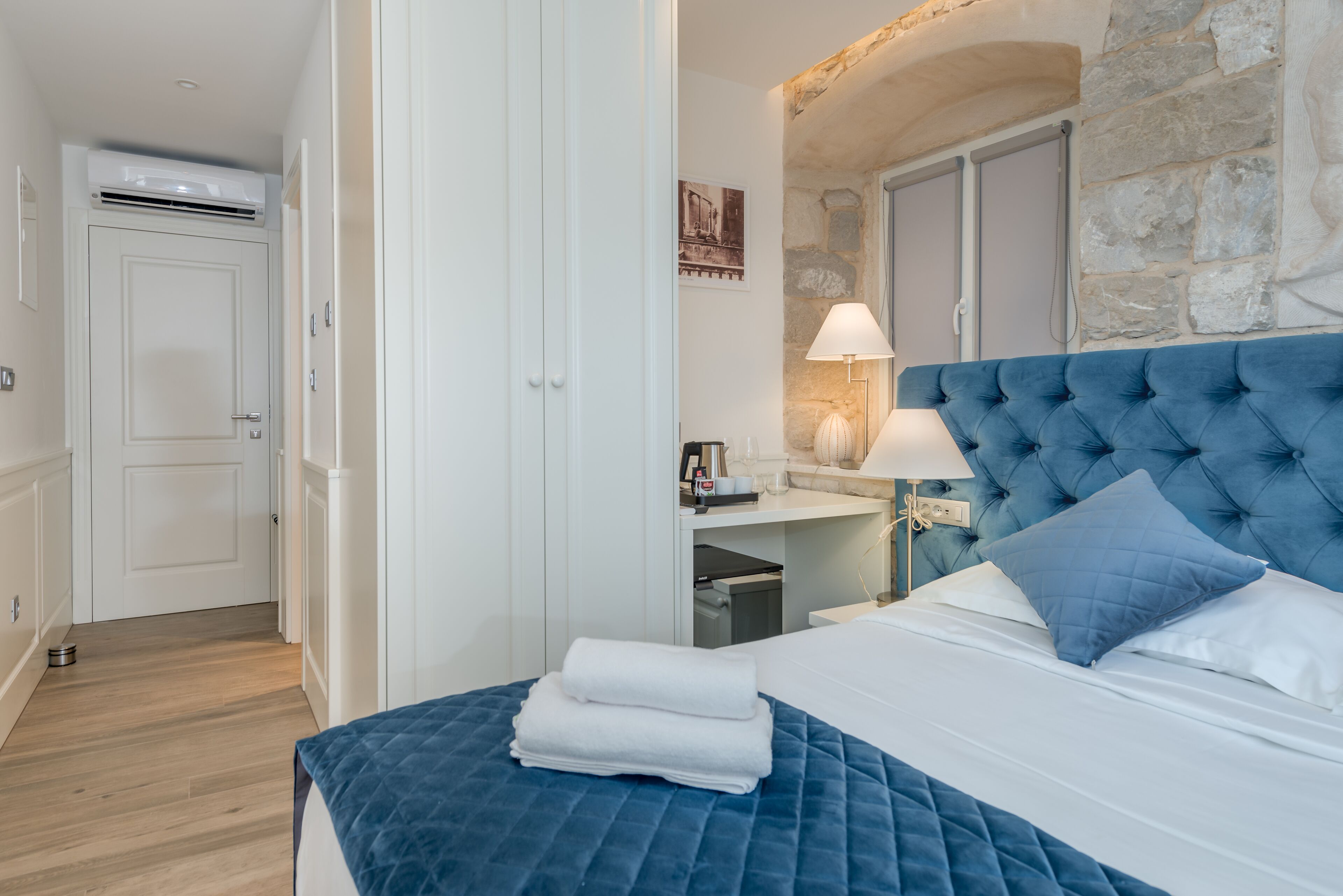Un dormitorio con un cabecero tapizado azul, una cama bien hecha con ropa blanca, una pared de acento de piedra y un armario blanco.
