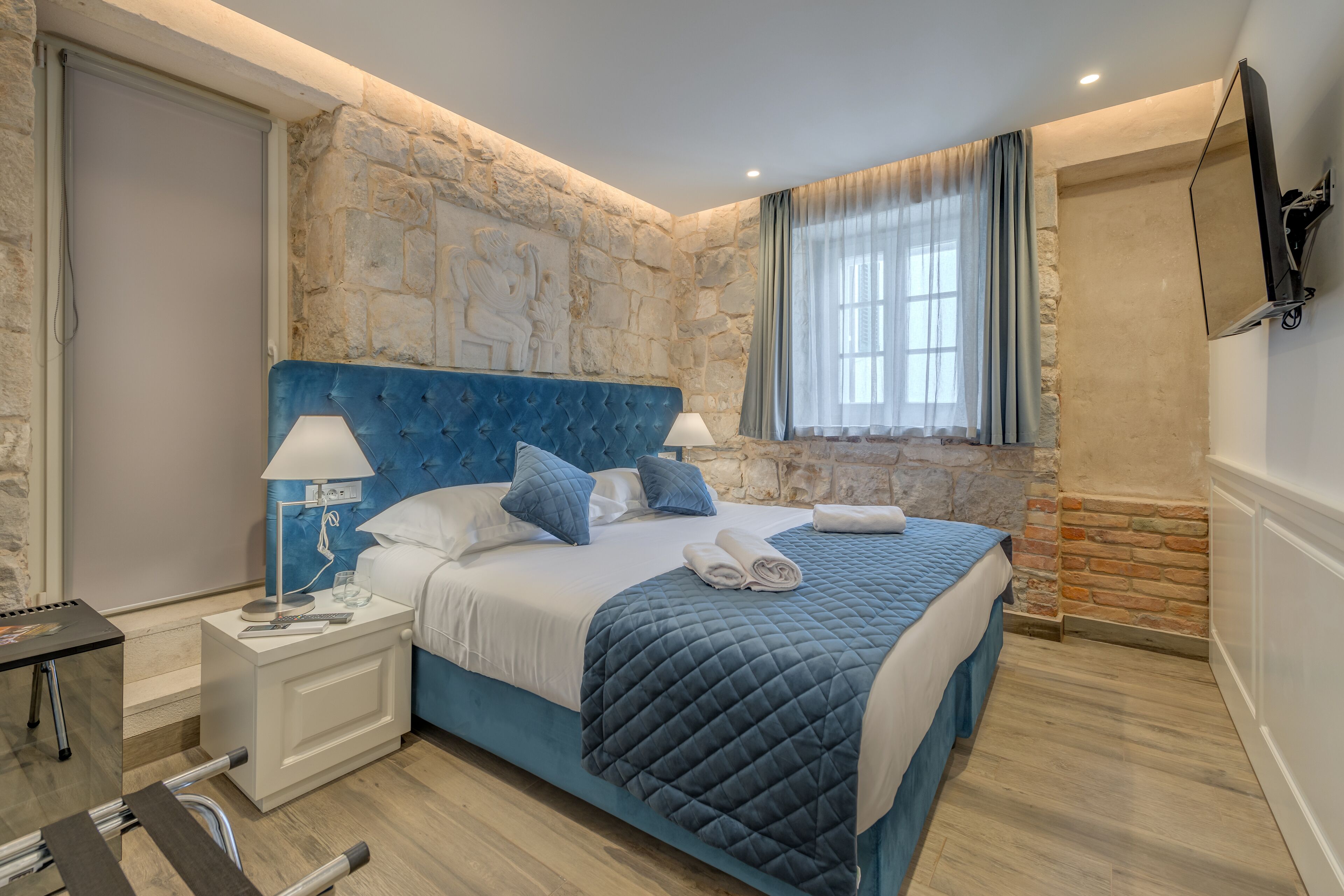 Un dormitorio con pared de piedra, cabecera tapizada en azul, cama con ropa blanca y una mesita de noche con lámpara.