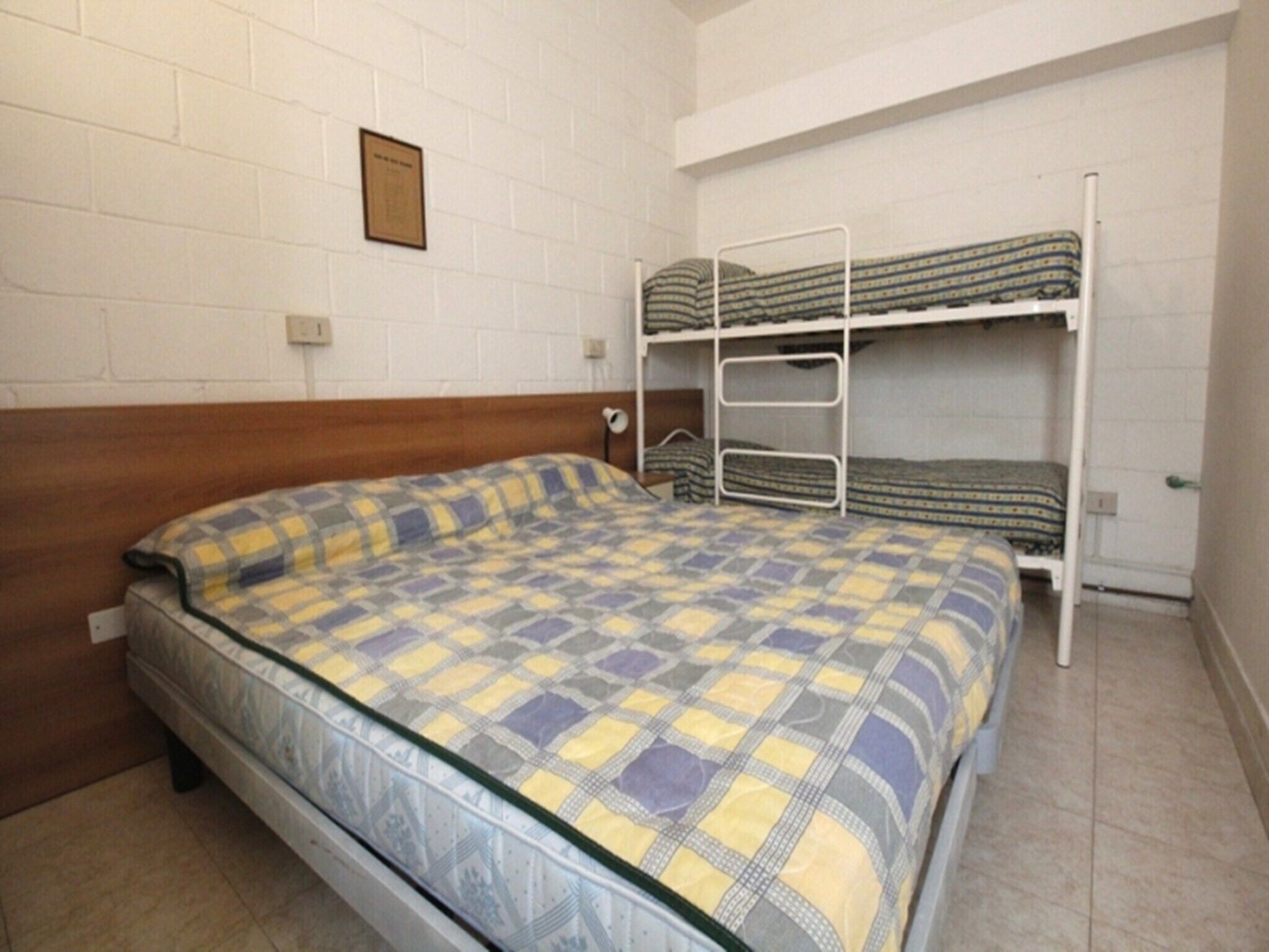 A single bed with a checkered bedspread, a wooden headboard, and a bunk bed with patterned bedding in the background.