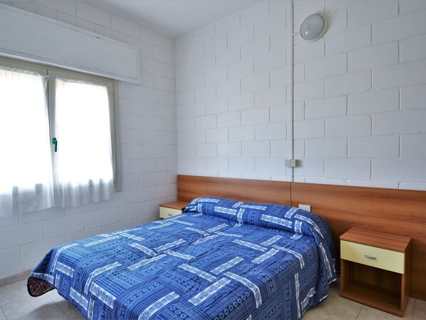 Une chambre à coucher avec une tête de lit en bois, un lit simple recouvert d’une courtepointe à motifs bleus, une table de chevet avec un tiroir et une fenêtre avec des rideaux.