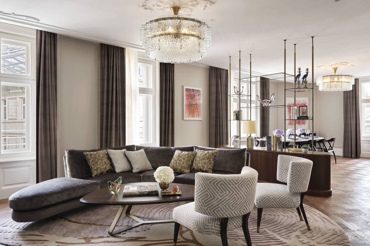 A spacious living room with a large sofa, two armchairs, a coffee table, and a chandelier. There is a dining area with a table and chairs in the background.