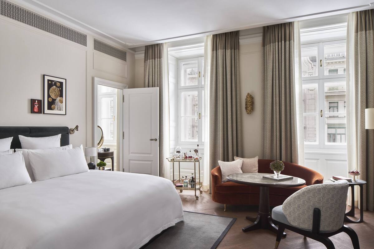 A hotel room with a large bed, a round table with a vase, a sofa, and a view of the city through large windows.