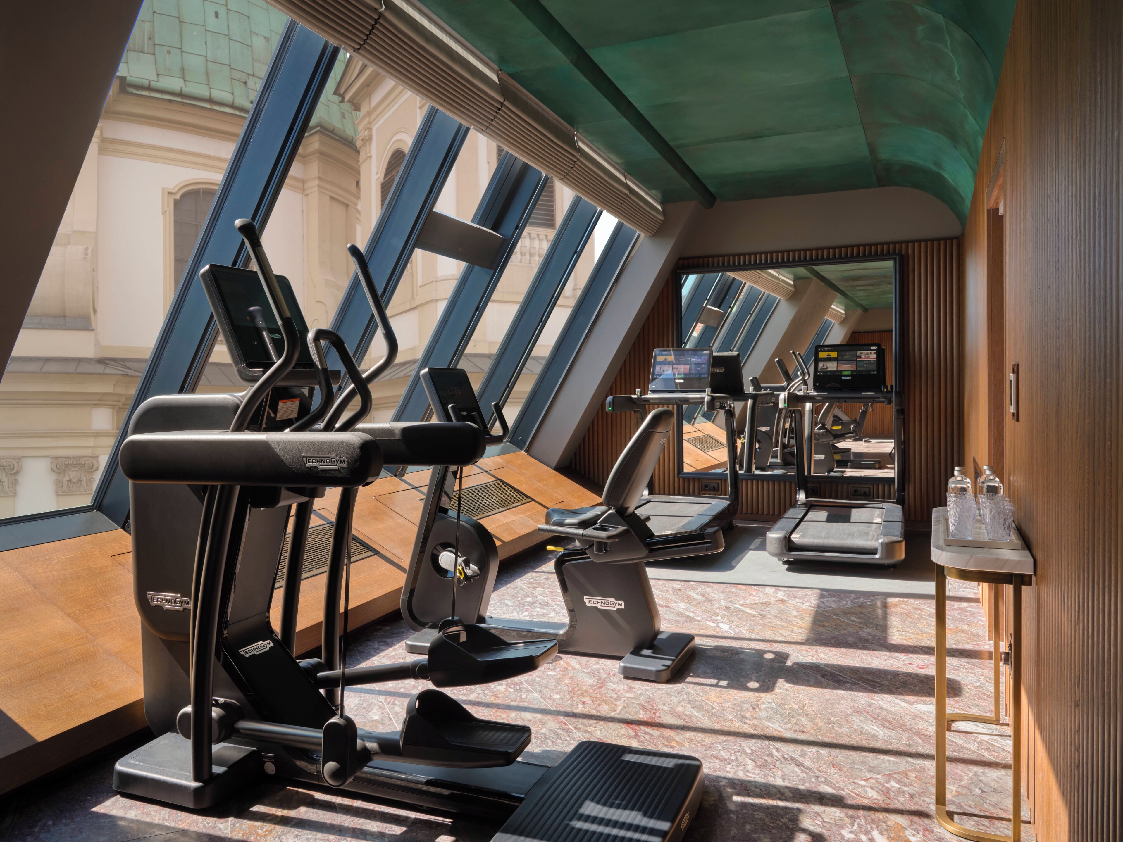 Ein Fitnessraum mit Trainingsgeräten, einem digitalen Bildschirm und großen Fenstern.