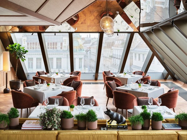 Un restaurante moderno con techos altos, amplios ventanales y vistas a edificios históricos. Las mesas están cubiertas con manteles blancos y sillas marrones.