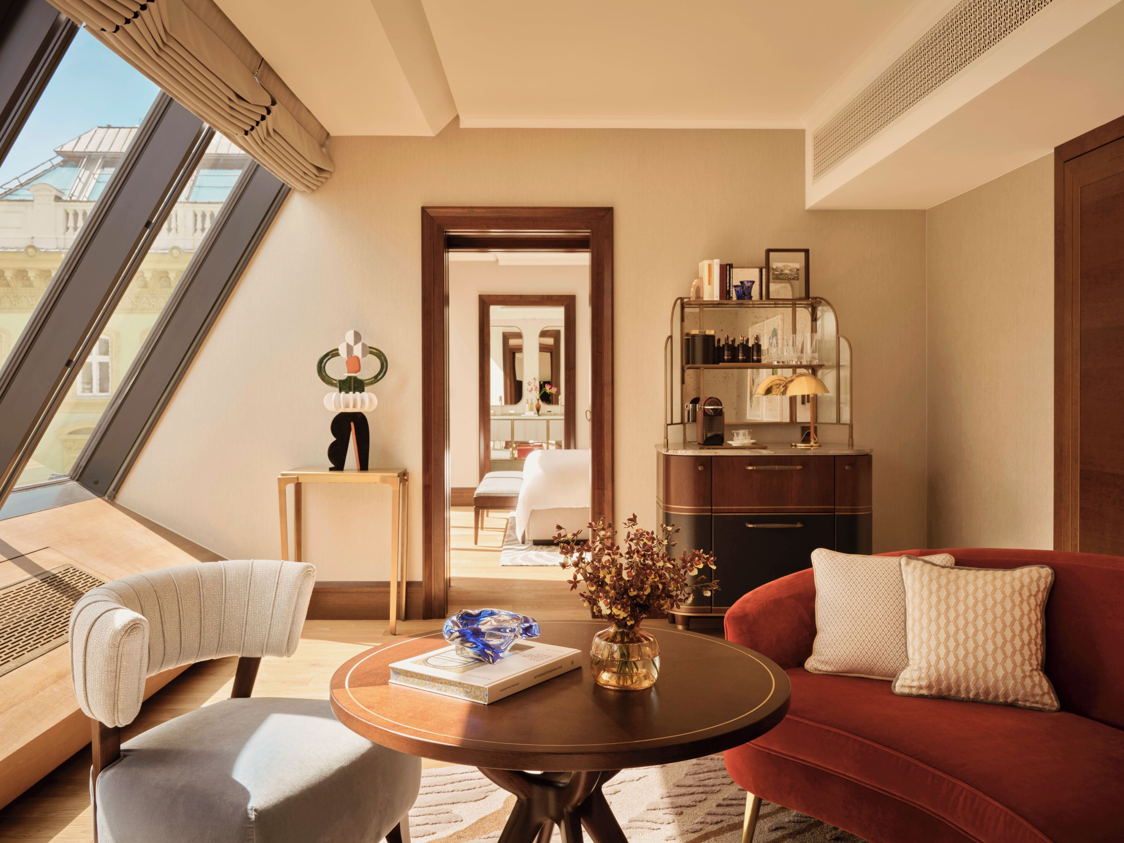 A modern hotel room with a large window, a red sofa, a round table with a book and vase, and a small side table with a decorative item.