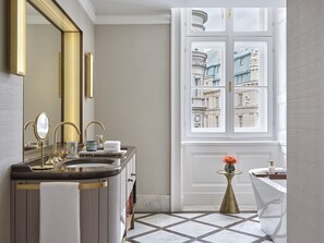 A modern bathroom with a double sink, a large mirror, and a view of historic buildings through a window. - Rosewood Vienna (Vienna)