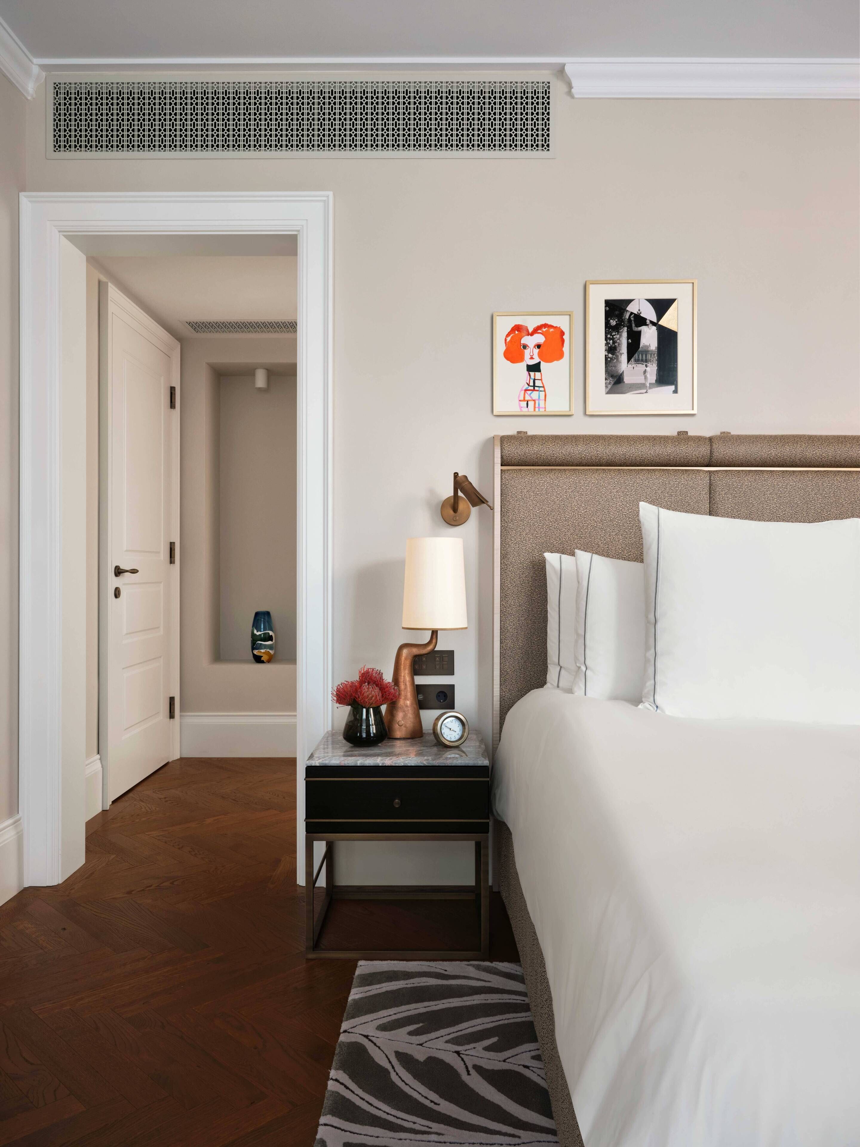 Habitación de hotel con cama, mesita de noche con lámpara, reloj y dos cuadros en la pared.