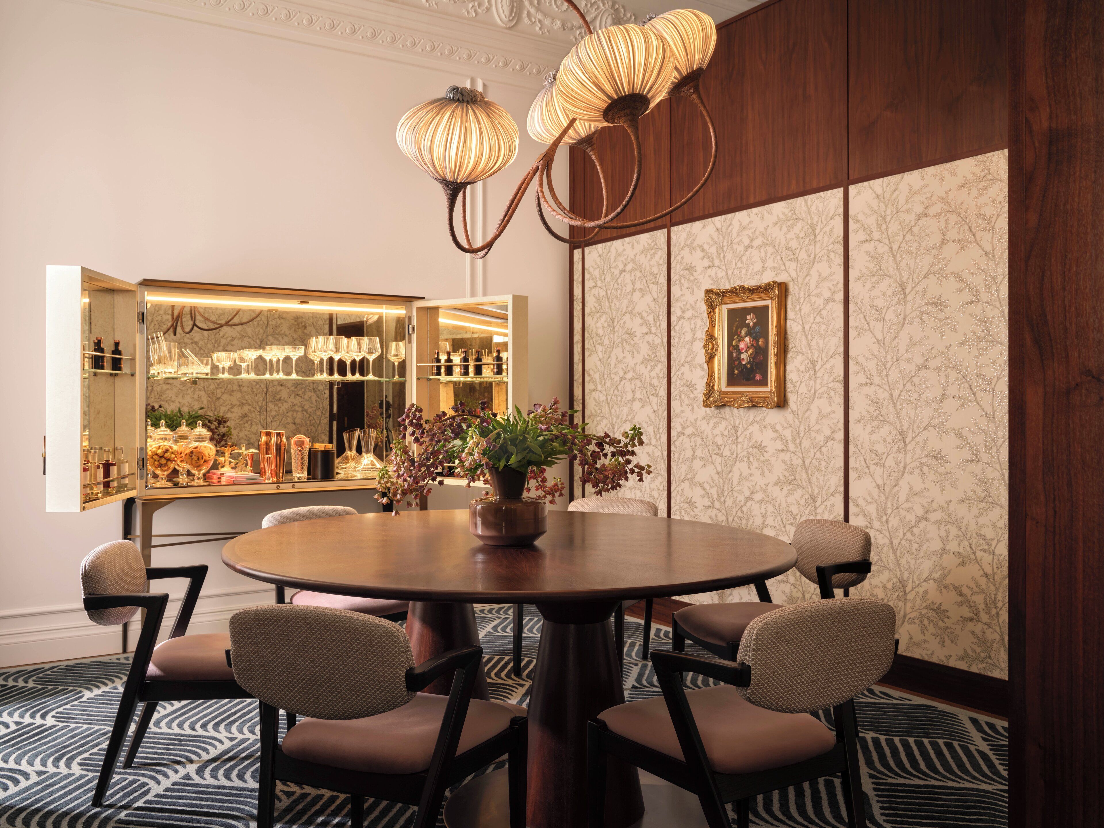A dining area with a round table, six chairs, a vase with flowers, a chandelier, and a display cabinet with glassware.
