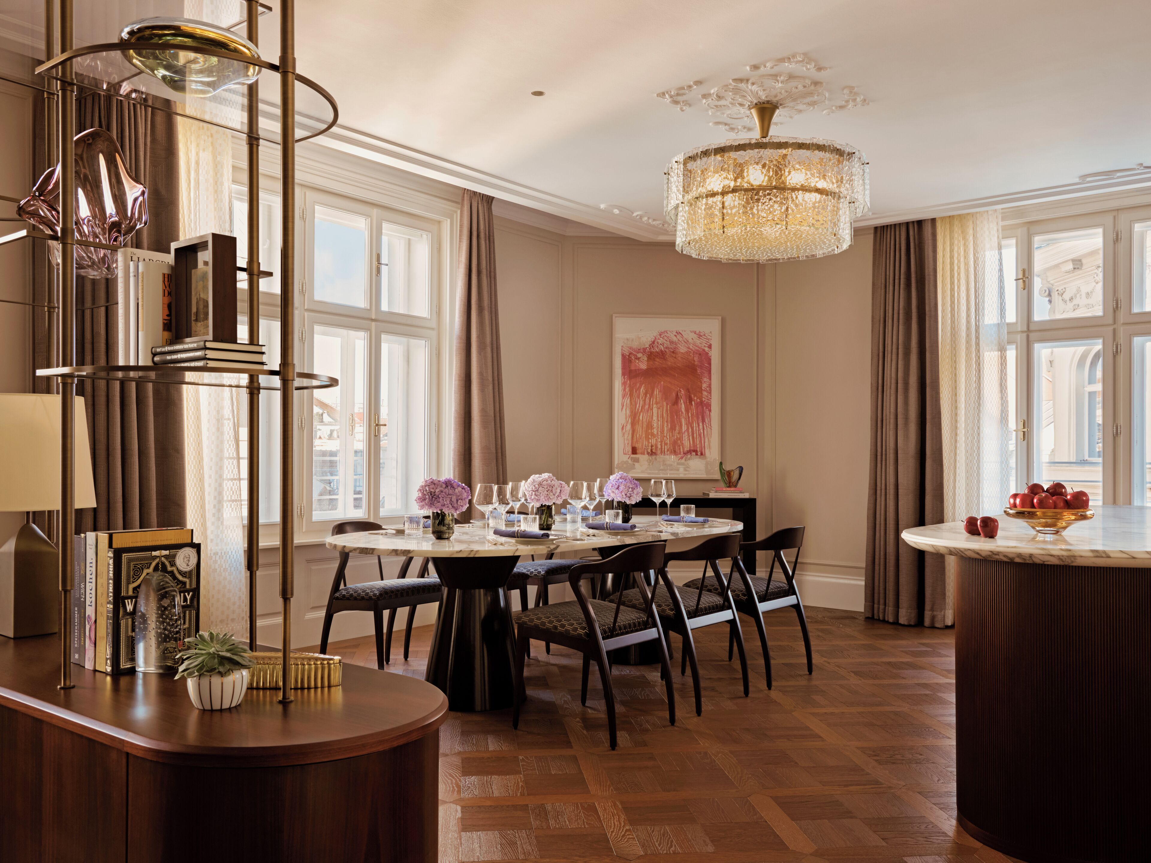 A dining area with a round table, chairs, a chandelier, and a view of the city through large windows.