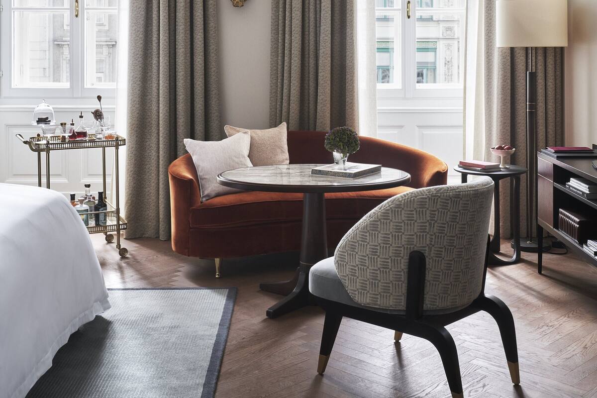 A hotel room with a bed, a sofa, a chair, a small table, and a lamp. There are curtains on the windows, and a decorative wall piece is visible.