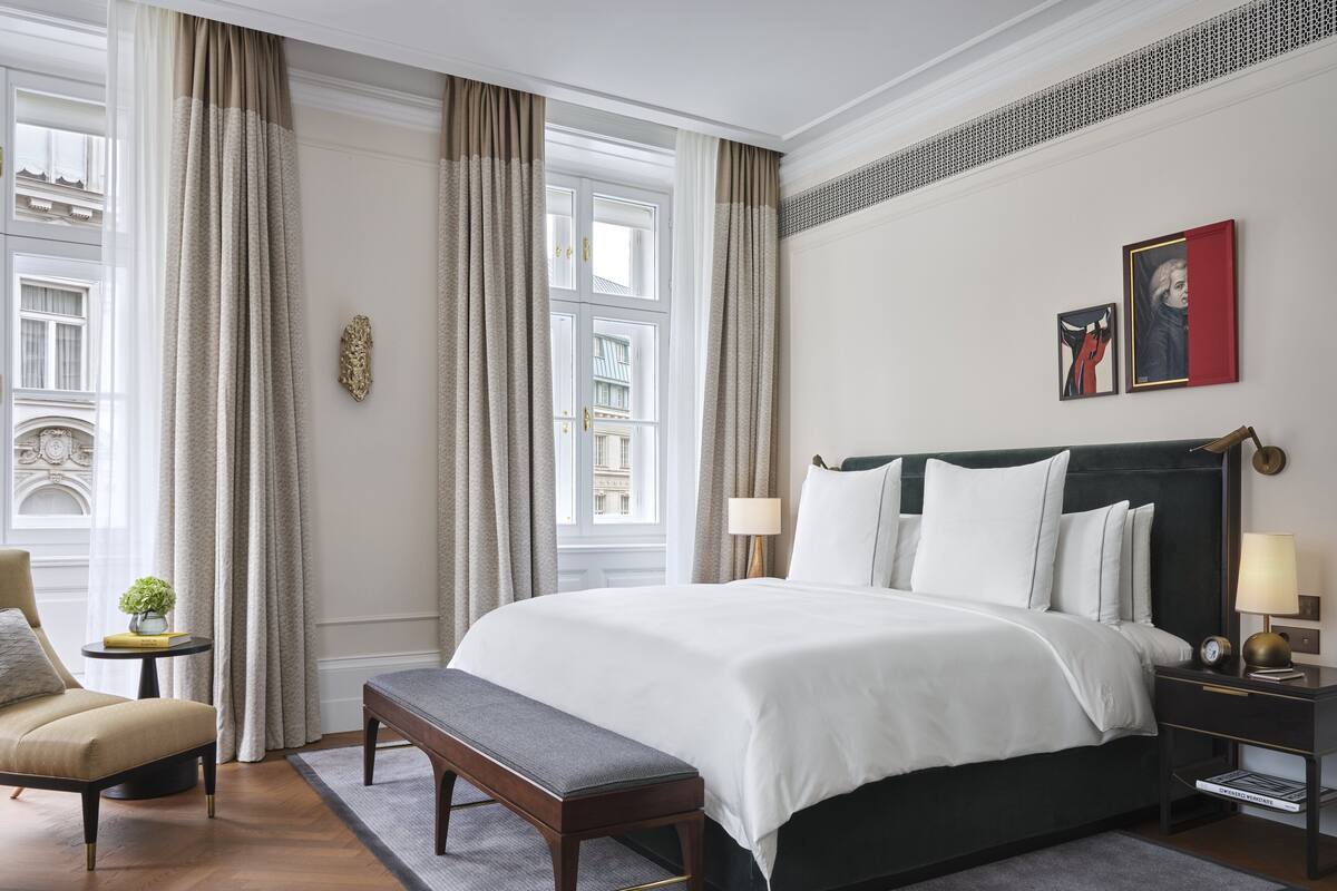 A hotel room with a large bed, a bench, a chair, a small table with a plant, and a view of the city through the window.