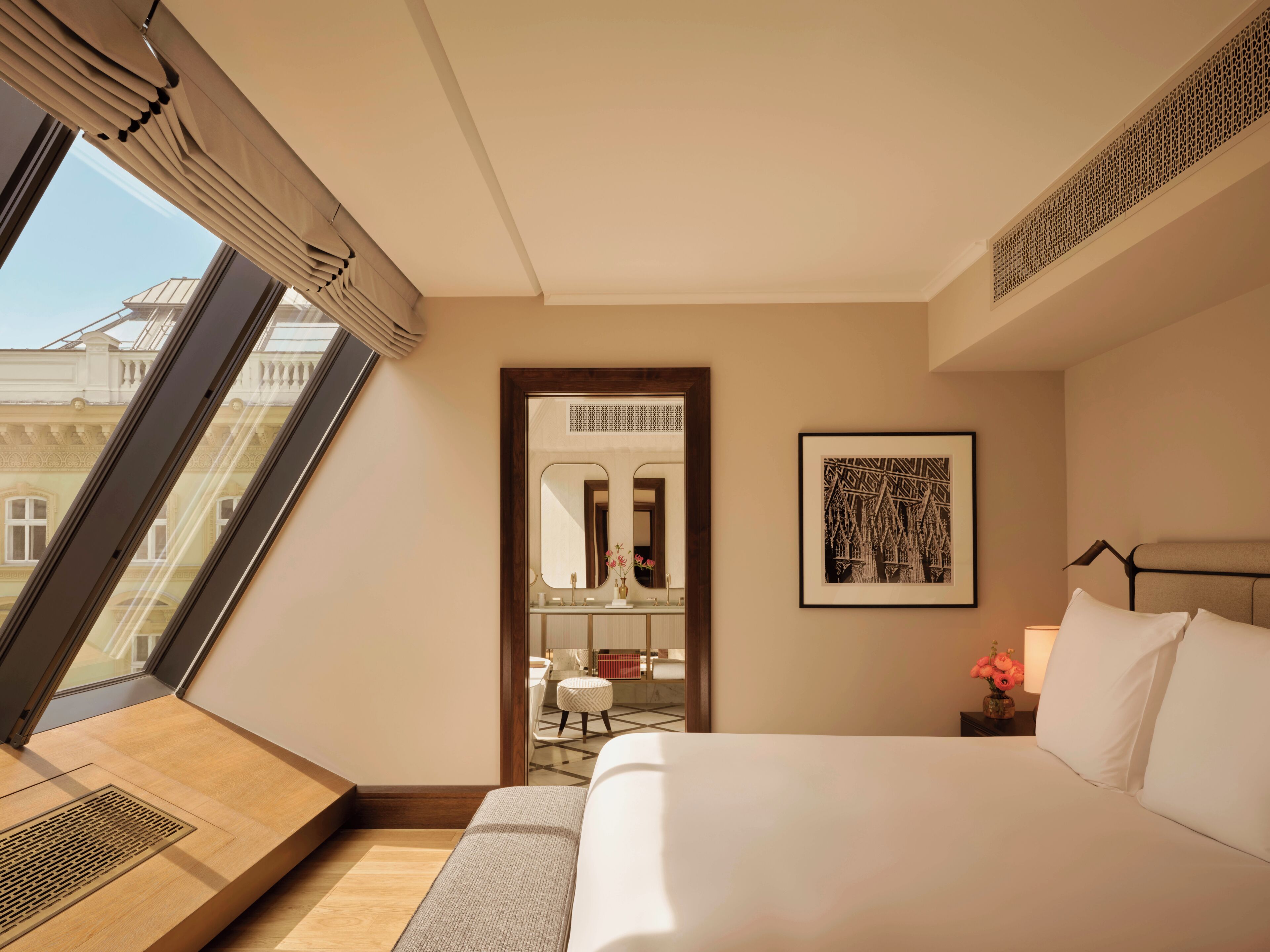 Una habitación de hotel con una cama grande, una obra de arte enmarcada, una mesita de noche y vistas a un edificio a través de una ventana.