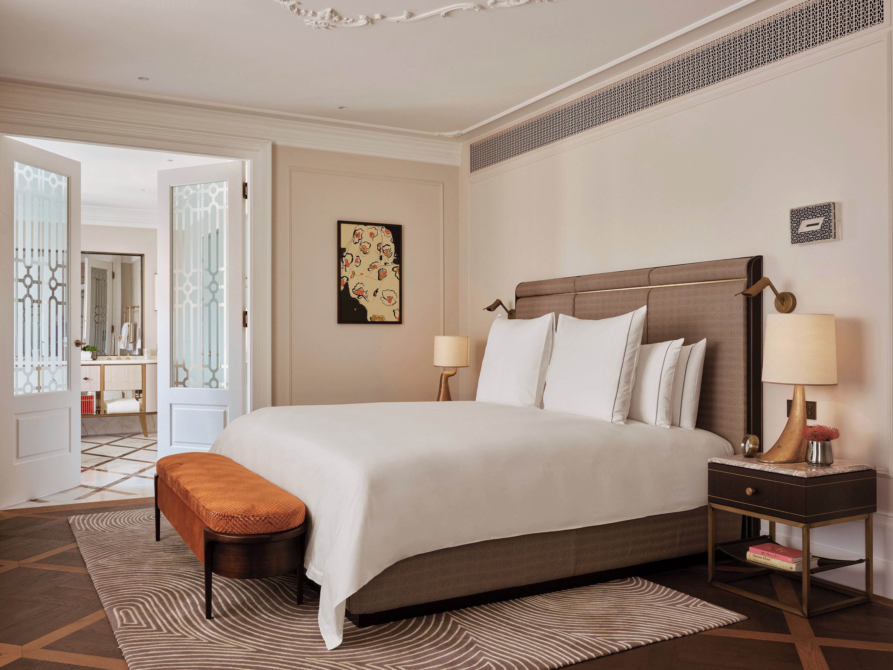 A hotel room with a large bed, a bedside table with a lamp, a bench, and a patterned rug.