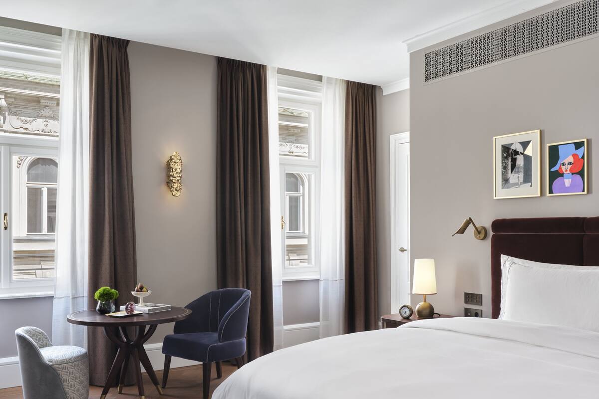 A hotel room with a large bed, a round table with two chairs, a small side table with a lamp, and two framed pictures on the wall.