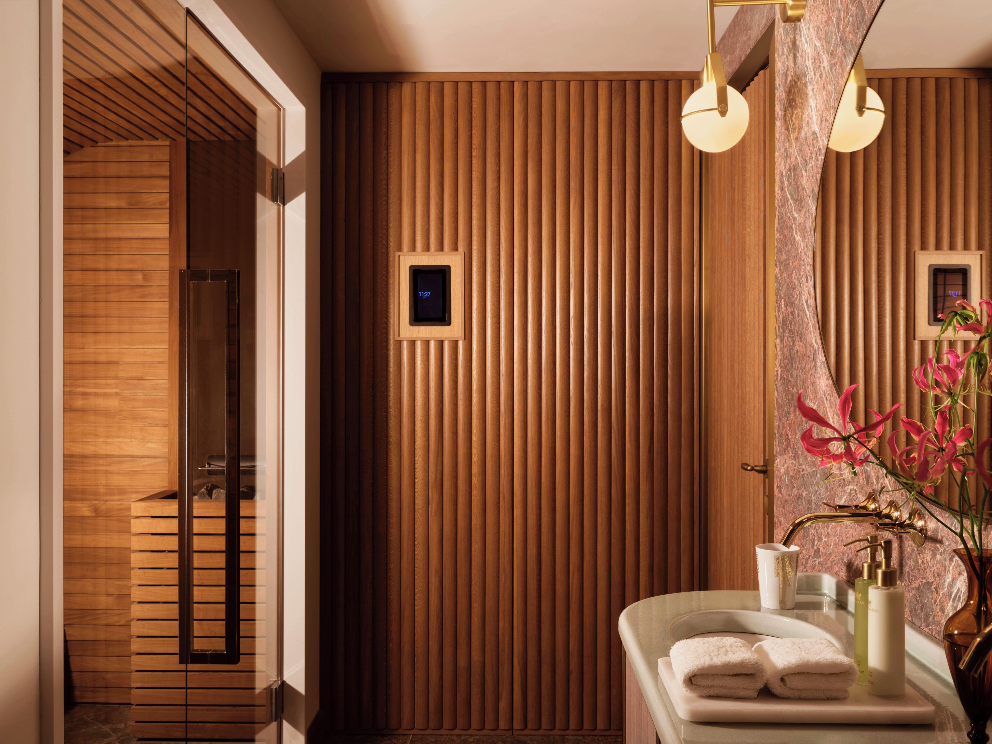 Un baño moderno con paneles de madera, un reloj digital, un tocador con espejo y un florero con flores.