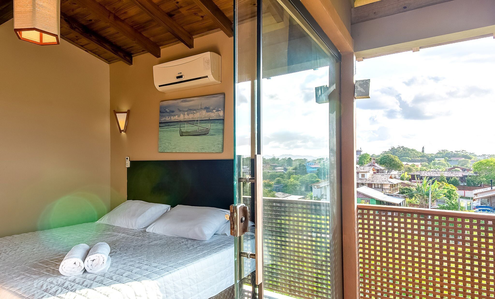 Un dormitorio con cama, dos almohadas, un cuadro, unidad de aire acondicionado y un balcón con vistas a un pueblo.