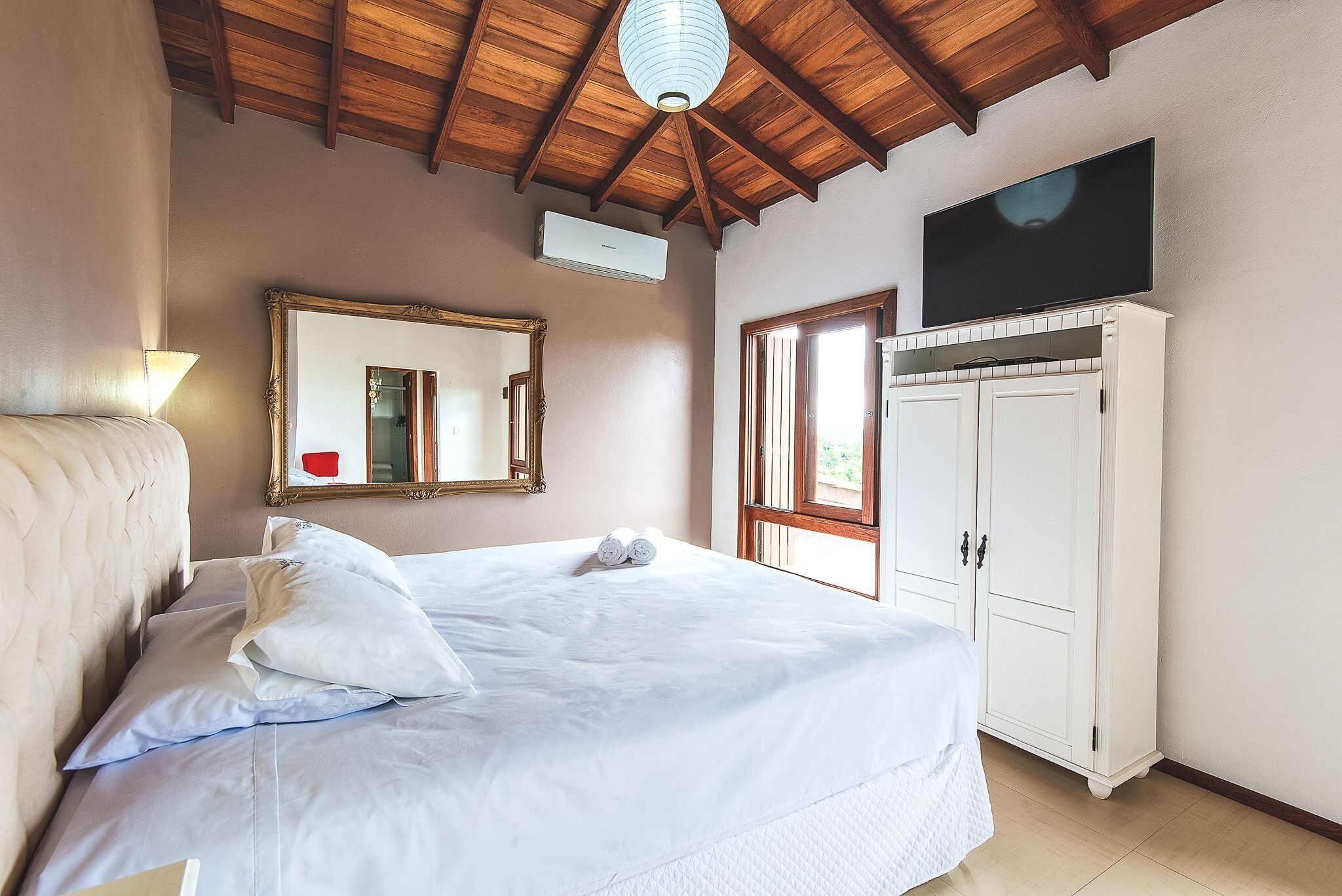 Uma cama bem arrumada com lençóis brancos, um espelho grande, uma televisão montada acima de um armário branco e um teto de madeira.