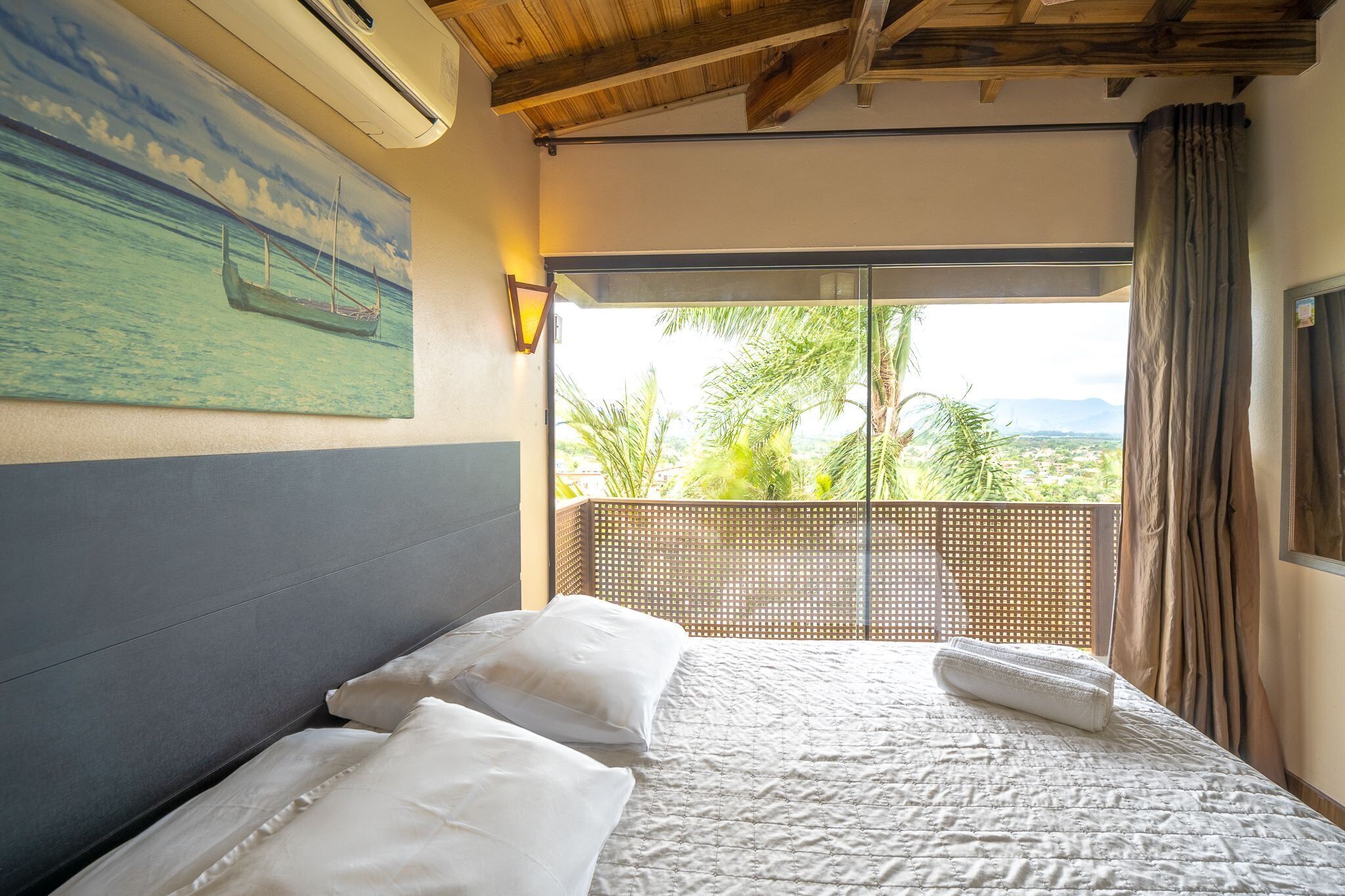 Quarto com cama, almofadas, um quadro de barco na parede e vista para palmeiras e montanhas pela janela.