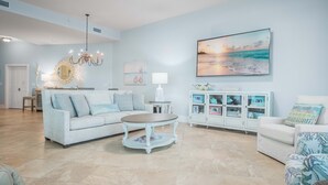 Ein geräumiges Wohnzimmer mit einem hellblauen Sofa, einem runden Couchtisch, einer weißen Vitrine mit Glastüren und einem großen Gemälde einer Strandansicht.